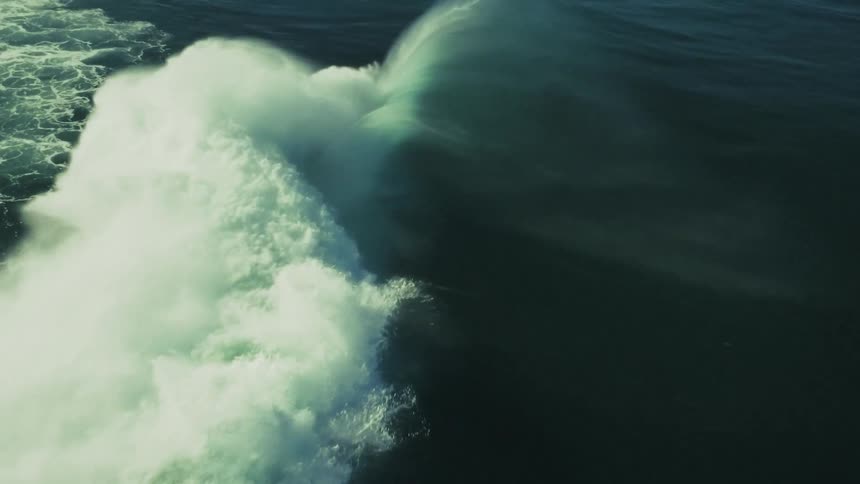 A dramatic aerial slow-motion shot captures a massive, powerful ocean wave as it forms, crests, and crashes