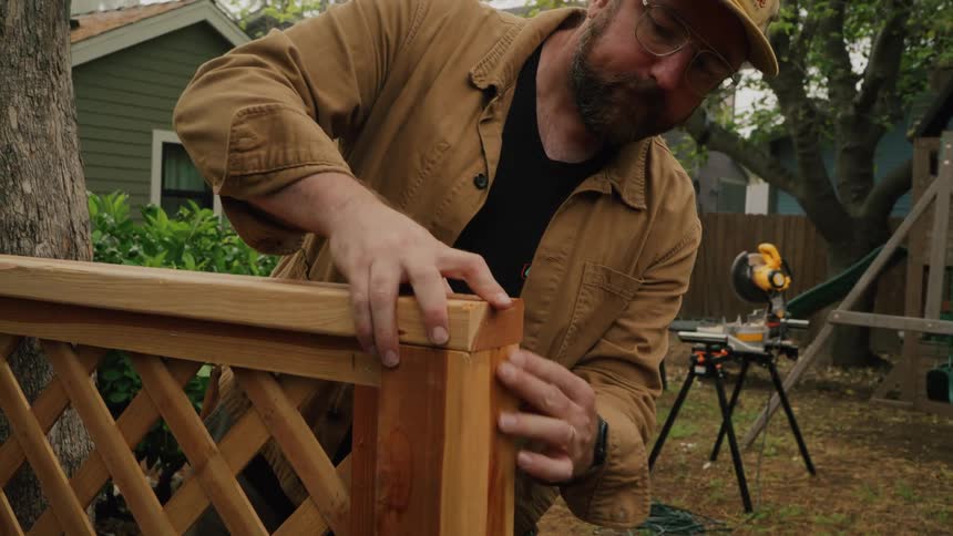 A bearded man wearing a cap and glasses meticulously checks the joints of a wooden fence he is building in his yard
