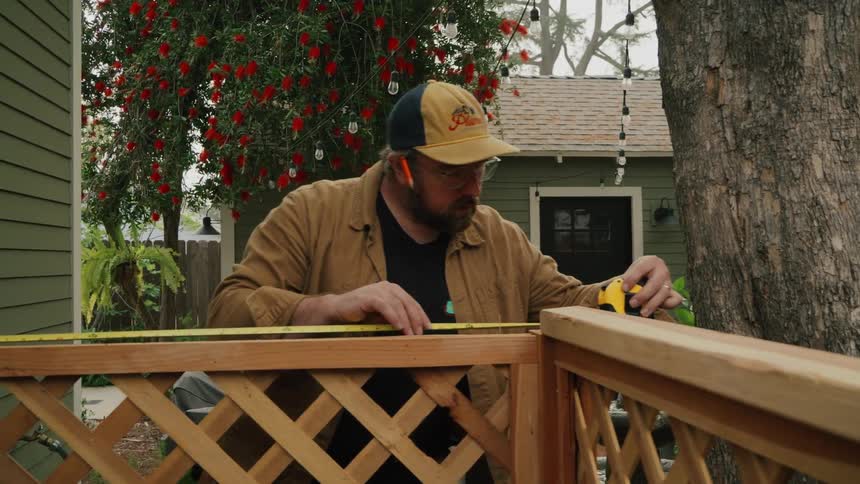 A man carefully measures a wooden lattice fence with a tape measure in a backyard setting