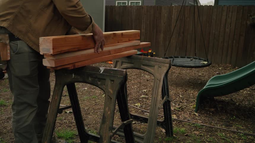A carpenter carefully carries a stack of heavy wooden beams and sets them on sawhorses in a backyard in slow motion