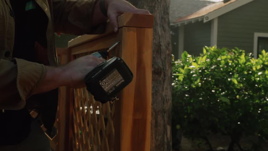 Slow motion tilt down shot of a focused man in a hat using a power drill to build a wooden fence in a sunny backyard