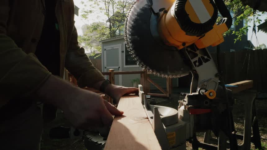 A woodworker measures and marks a piece of wood before making a cut with a large miter saw in a backyard
