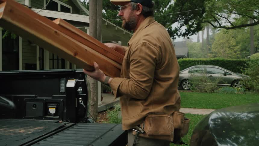 A man wearing a tool belt unloads wooden beams from the back of a truck in slow motion