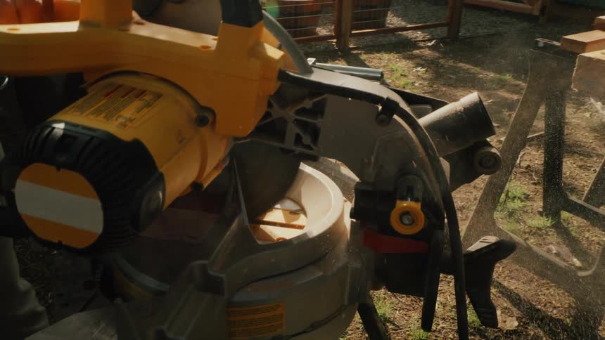 A close-up slow-motion shot of a miter saw cutting through a piece of wood, sending sawdust flying through the air in a sunny backyard