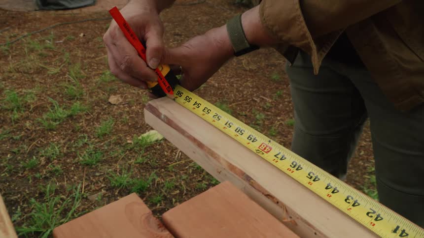 A focused woodworker in his backyard carefully measures a plank of wood with a tape measure and marks it with a pencil