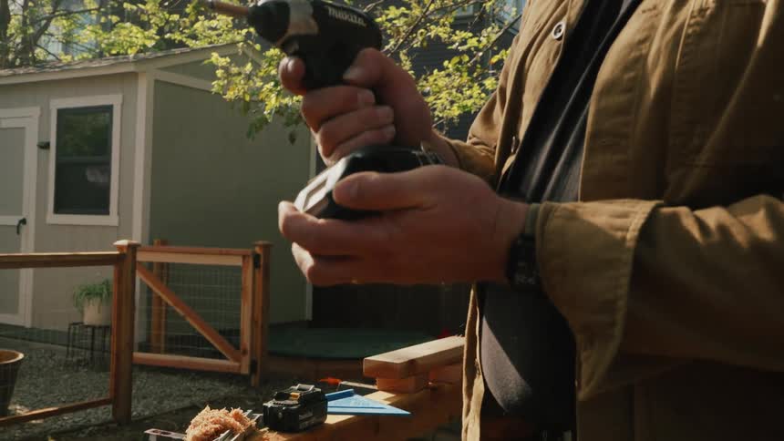 A man in a brown work jacket puts a battery into a cordless power drill in his backyard in slow motion