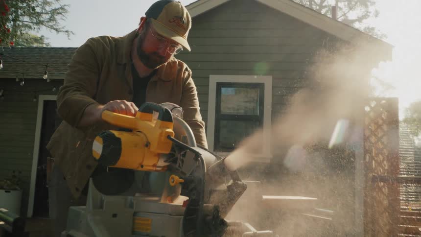 Slow motion shot of a carpenter using a miter saw to cut wood in a sunny backyard, with sawdust particles suspended in the air