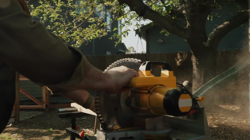 A professional carpenter makes a precise cut on a wood plank with a miter saw in his backyard on a sunny day, captured in slow motion