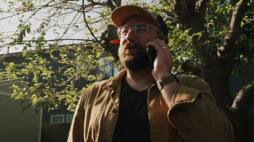 A man with a beard, glasses, and a hat talks on his smartphone in a sunny backyard