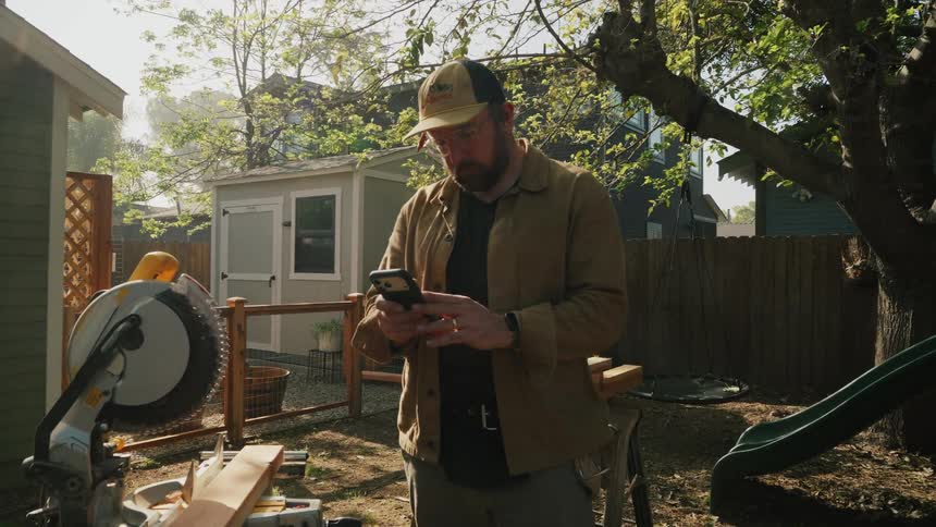 A focused carpenter takes a moment to check his smartphone for instructions while working on a project in his sunny backyard