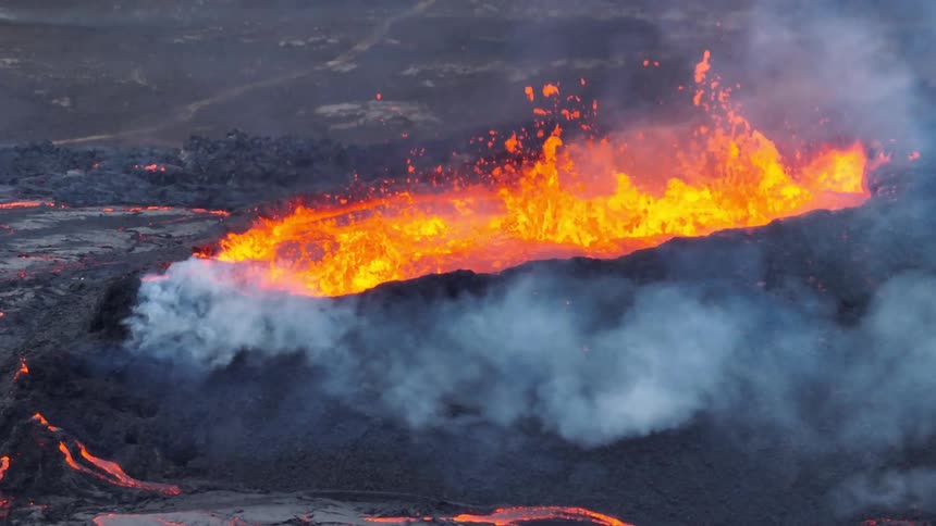 A dramatic aerial view zooms out from a powerful volcanic eruption, revealing fiery lava spewing from the crater and flowing across the dark landscape