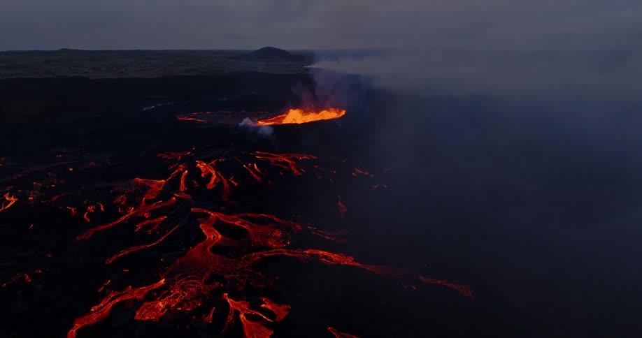 A powerful aerial shot captures a fissure volcano erupting at night, with rivers of molten lava illuminating the dark, smokey landscape