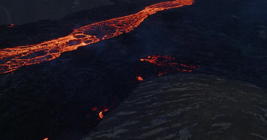 A dramatic aerial shot reveals a glowing river of lava flowing from an erupting volcano through a dark, desolate landscape at dusk