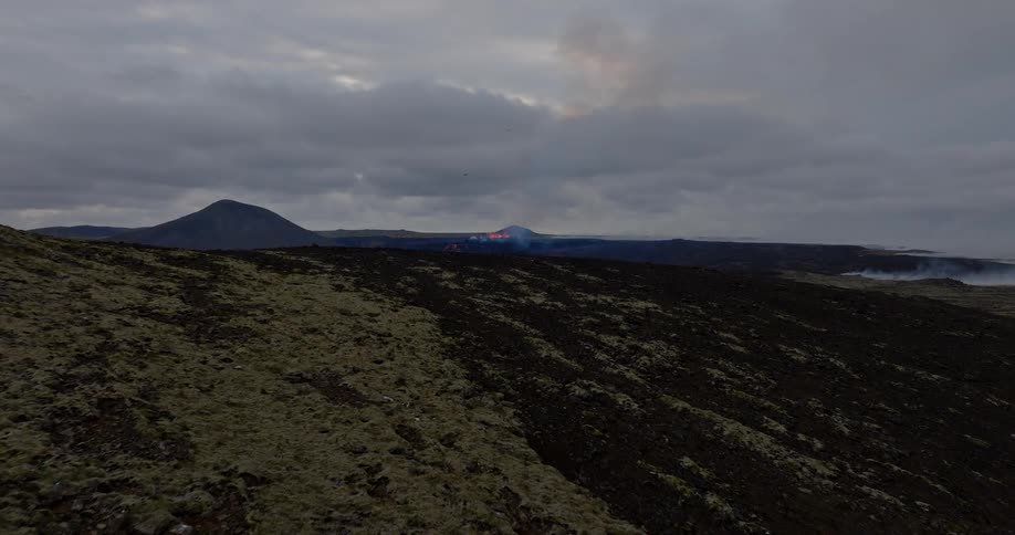 An aerial drone shot flies over a mossy hill to reveal the Fagradalsfjall volcanic eruption in Iceland under a cloudy sky