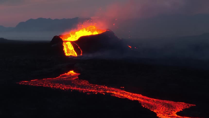 A volcano erupts with fiery lava spewing from its crater and a glowing river of molten rock flowing across the dark landscape at dusk