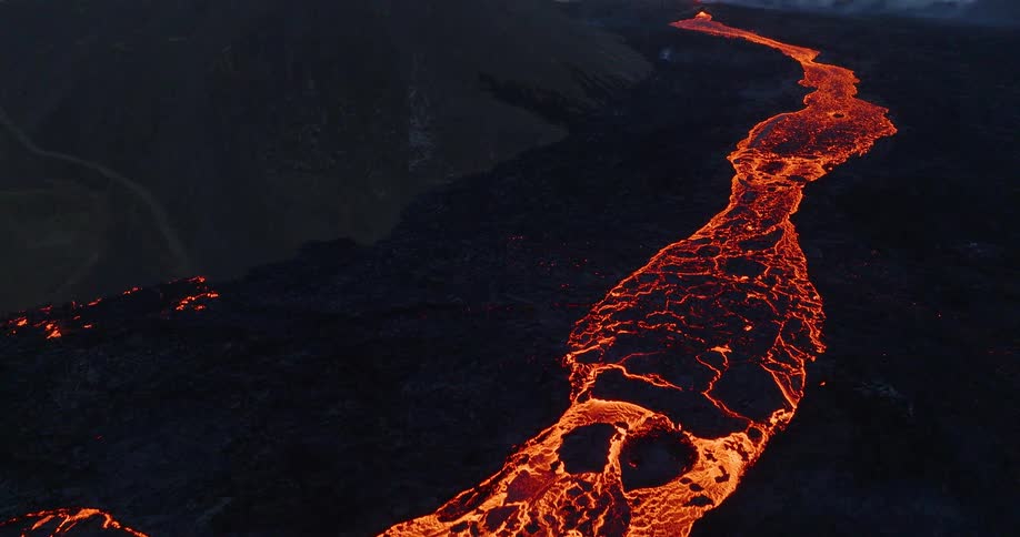 Dramatic aerial shot pulls back to reveal a massive river of molten lava flowing from an erupting volcano at dusk