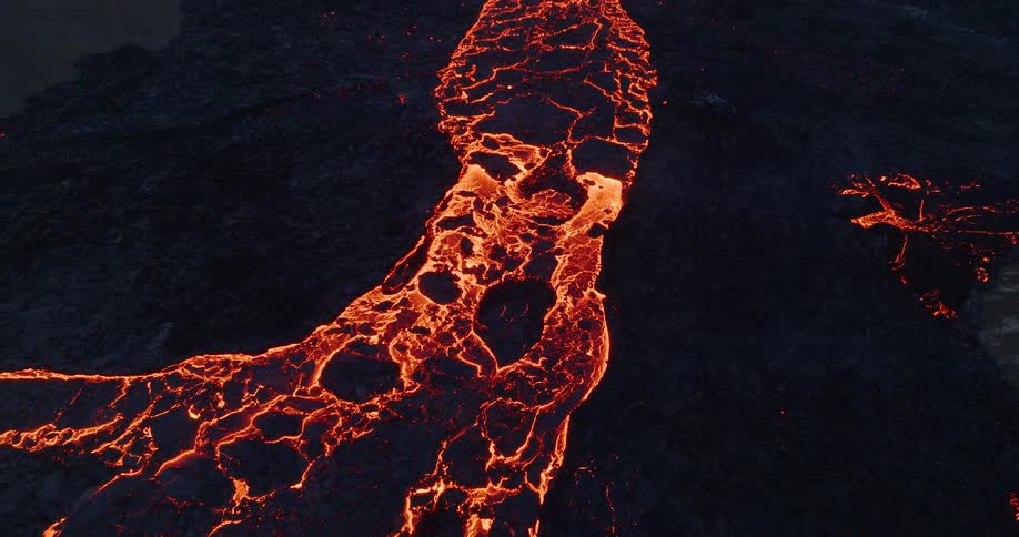 An aerial drone shot pulls back from a glowing river of molten lava to reveal a spectacular volcanic eruption at dusk
