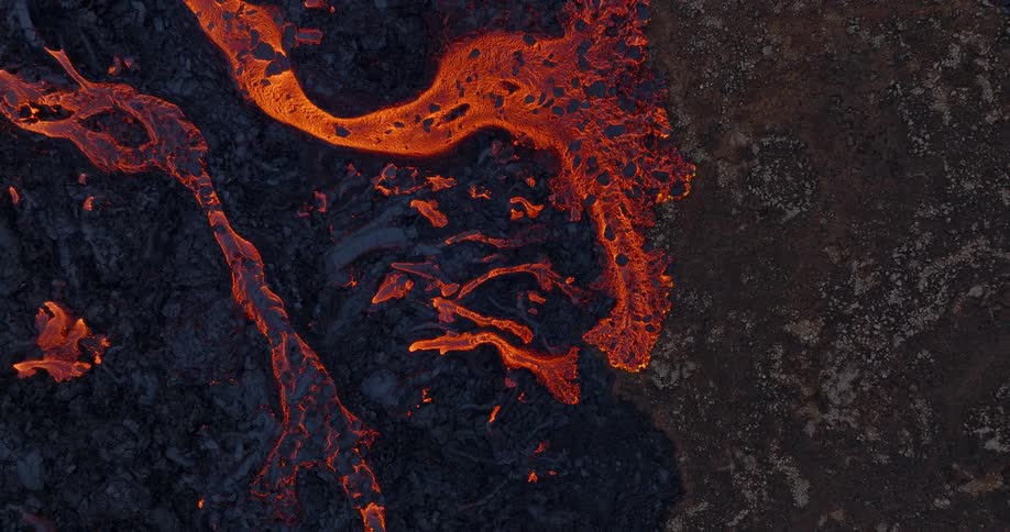An overhead aerial view captures the mesmerizing flow of molten lava as it slowly spreads across a field of dark, cooled volcanic rock
