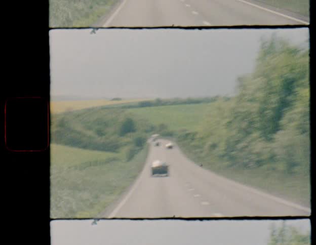 A shaky, blurry POV shot from a moving car follows a convoy of classic cars down a scenic rural highway, captured on vintage film