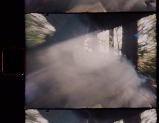 Grainy vintage film captures a person tending to a smoky campfire under a rustic shelter in the woods