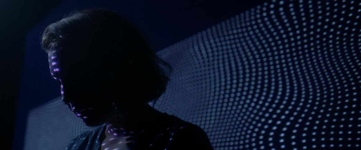 A young woman sits in a dark a white light showing a pattern on her face and wall