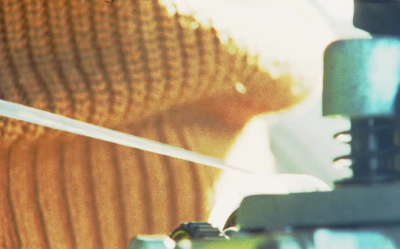 A vintage, grainy close-up shows a single white thread being processed through an industrial machine