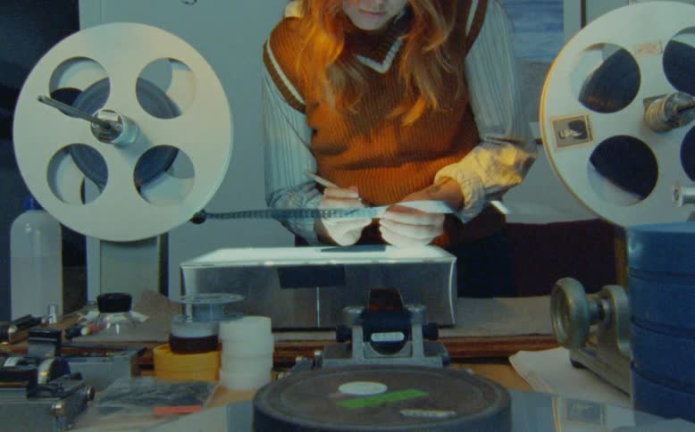 A film editor meticulously inspects a strip of film on a vintage editing bench with a lightbox