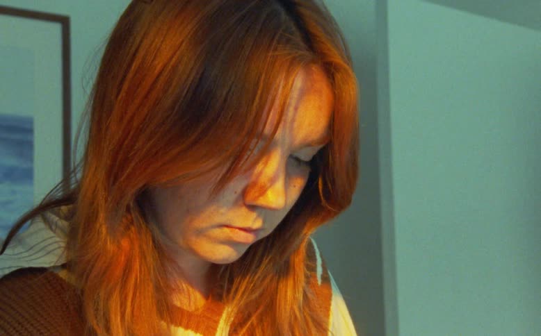 A close-up shot of a young woman with red hair, bathed in warm golden light, looking down thoughtfully