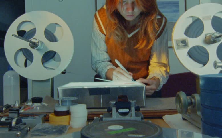 An artist meticulously hand-paints and animates directly onto a celluloid film strip using a vintage editing station with a lightbox