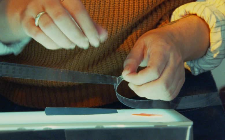 A film archivist carefully cleans and repairs a vintage film strip using tweezers over a glowing light table