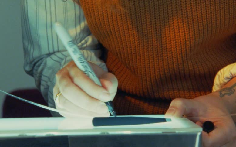 An artist with a tattoo carefully traces lines with a black marker and ruler on a lightbox