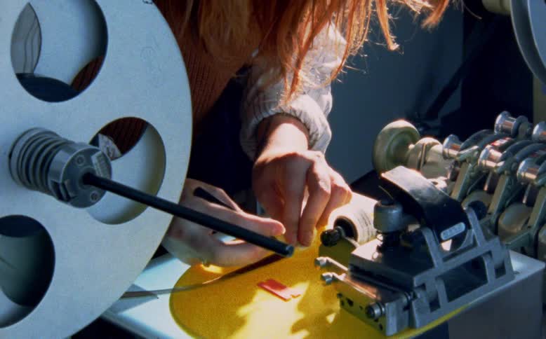 A close-up shot pulls back to reveal a filmmaker meticulously splicing together strips of film on a vintage editing machine