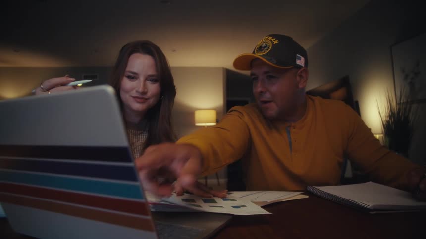 A veteran and his partner work together at their dining table late into the night, discussing charts and pointing at a laptop screen