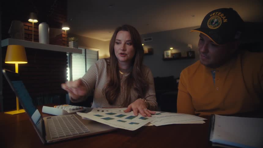 A military veteran and his wife sit at their dining table at night, seriously discussing their finances and reviewing charts on paper and a laptop