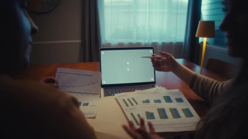 Two colleagues analyze financial data on charts and a green screen laptop while working late at a dining room table