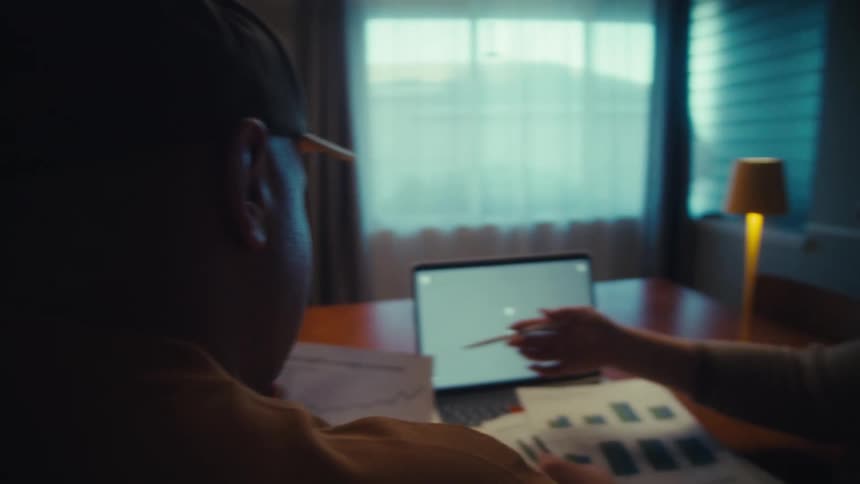A man and a woman review business charts and graphs on a laptop with a green screen during a meeting in a dimly lit room