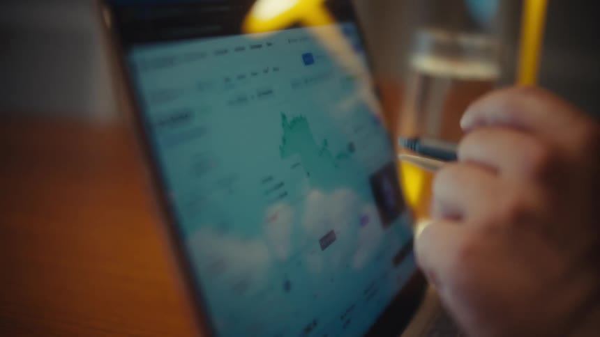 A financial analyst uses a stylus to closely examine a rising stock market graph on a laptop screen in a dimly lit room