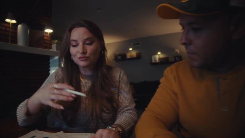 A focused woman explains charts and data on papers to her veteran husband while sitting at a table in their home at night