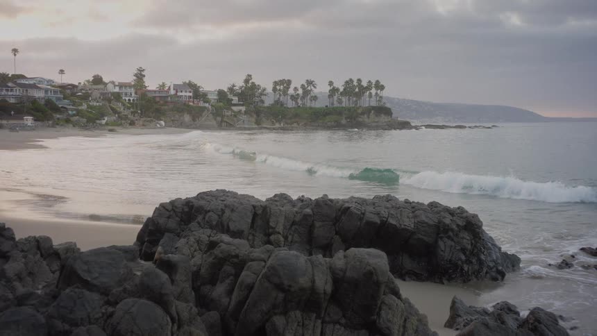 Calm ocean waves roll in slow motion onto a rocky shoreline with a coastal town in the background on an overcast day