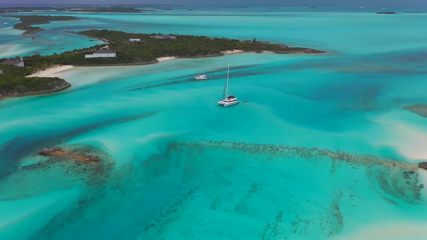 An aerial view captures a catamaran anchored in the serene, shallow turquoise waters surrounding a tropical island
