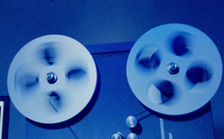 A close up shot of a vintage reel-to-reel tape recorder with the reels spinning quickly in a cool blue light