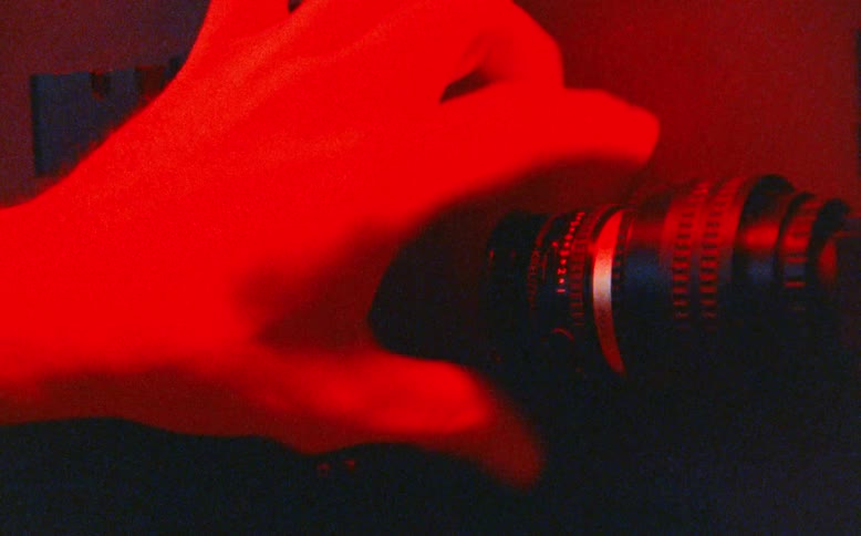 A technician's hands adjust the focus on a camera lens in a lab under shifting green, blue, and red lights
