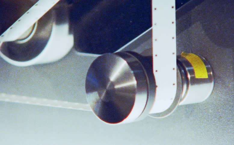 A close-up shot captures the intricate movement of a sprocketed film strip running through the metal rollers of a film transport mechanism