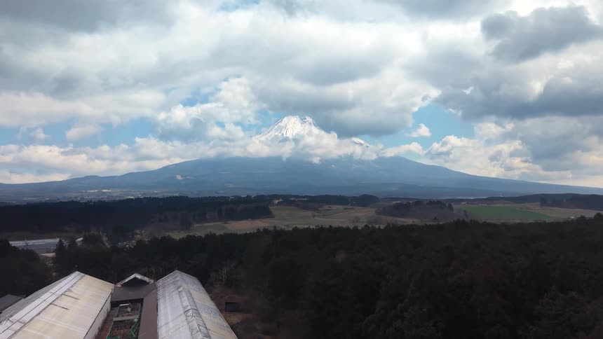 A slow aerial ascent reveals the majestic snow-capped Mount Fuji rising above a dense forest and the Japanese countryside under a cloudy sky