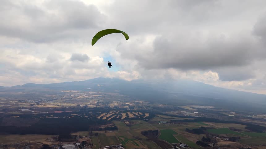 A paraglider soars majestically over a vast rural landscape with a large mountain looming in the background under a cloudy sky