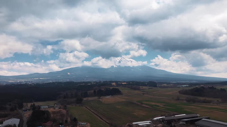 A slow aerial pullback reveals a rural Japanese village nestled in a valley with a majestic, snow-capped mountain under a cloudy sky