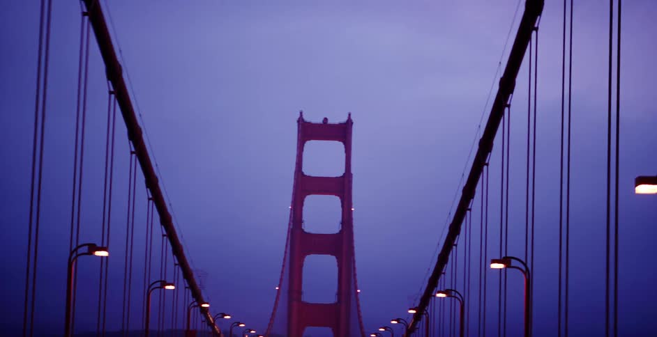 A moody pull-back shot from a moving vehicle reveals the iconic tower of the Golden Gate Bridge at twilight as the road lights illuminate the path forward