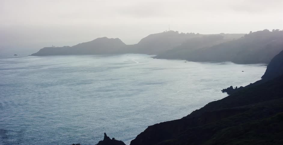 An atmospheric tracking shot reveals a serene and foggy coastline meeting the calm ocean waters on an overcast day