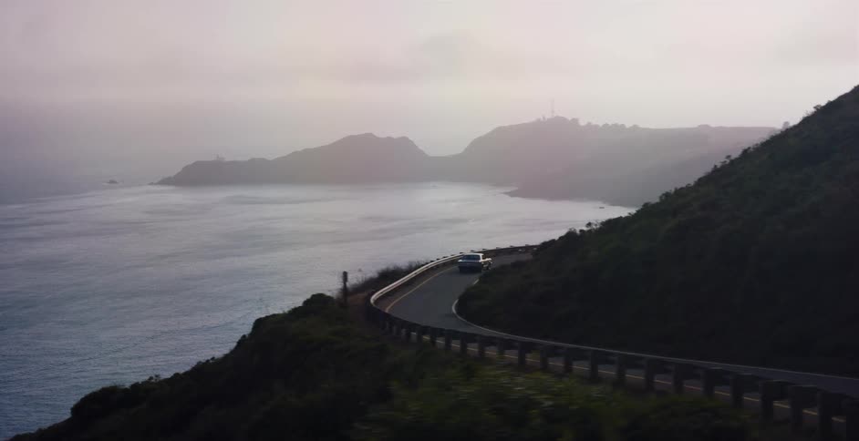 An aerial view shows a car driving on a winding coastal road next to the ocean on a foggy, overcast day