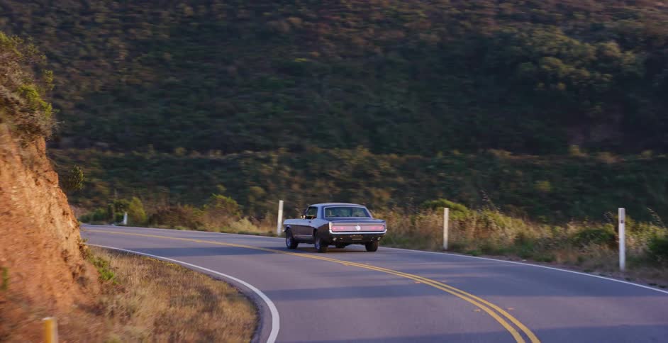 Tracking shot of a classic American muscle car driving along a winding coastal road at sunset